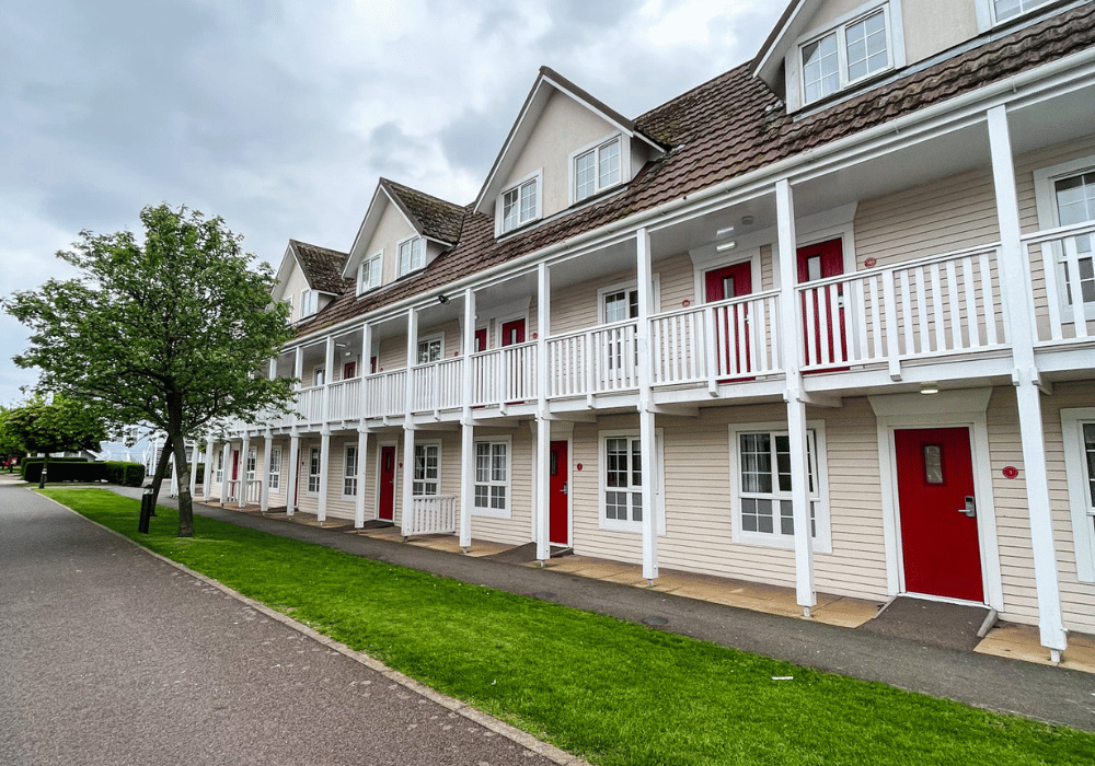 1 Butlins Fairground Apartments A row of two-story beige apartments with white trim and red doors. Each unit has a balcony and a small porch. A tree and green lawn line the pathway in front of the buildings under a cloudy sky.