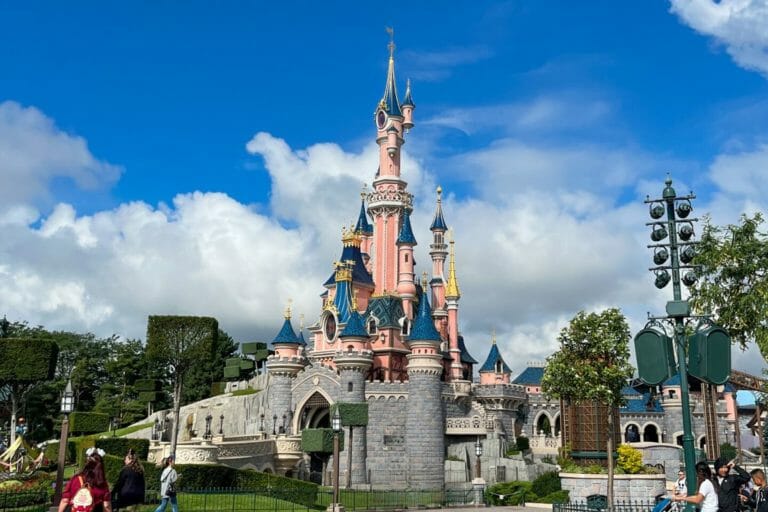 The iconic Sleeping Beauty Castle at Disneyland Paris, with its pink and blue towers, surrounded by manicured gardens and visitors, under a bright blue sky with scattered clouds.
