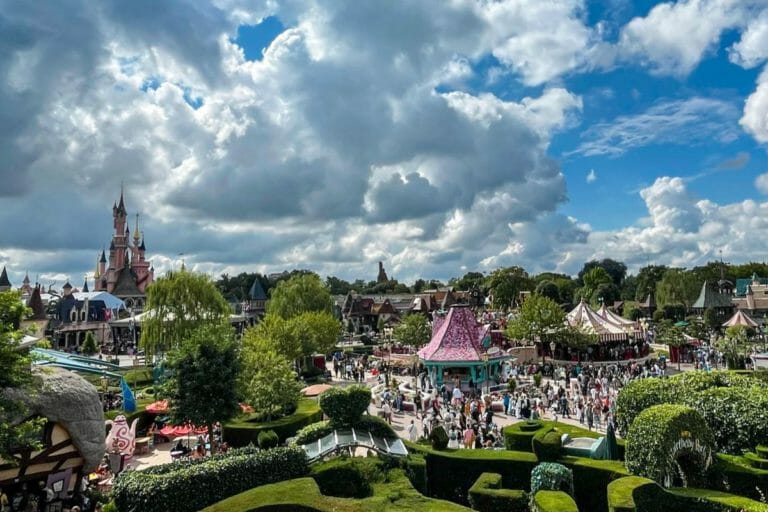 A wide view of Disneyland Paris filled with colorful buildings, various attractions, and Sleeping Beauty's Castle in the distance, under a partly cloudy sky, with crowds of visitors.