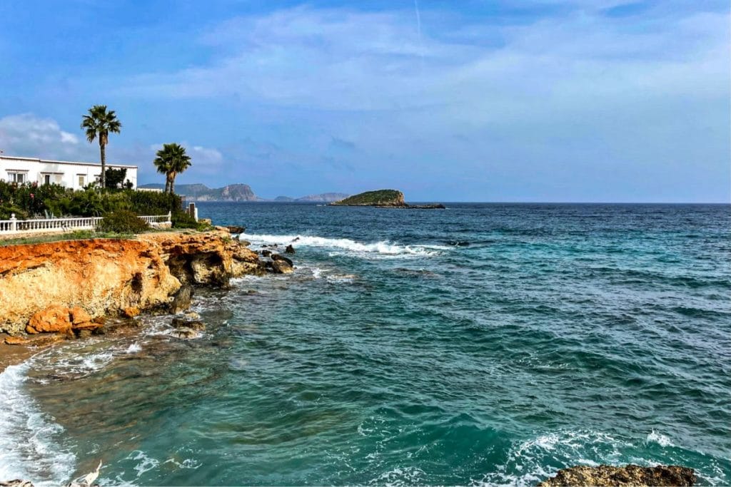 1 Ibiza Coastline The image shows a rocky coastline of Ibiza with turquoise waves crashing against it. On the left, there are palm trees and a white building with greenery. The sky is clear with distant islands visible.