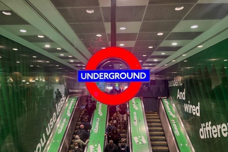 An entrance to the London Underground with a large red and blue roundel sign that reads 'Underground.' The entrance features escalators leading down, with passengers visible on them.
