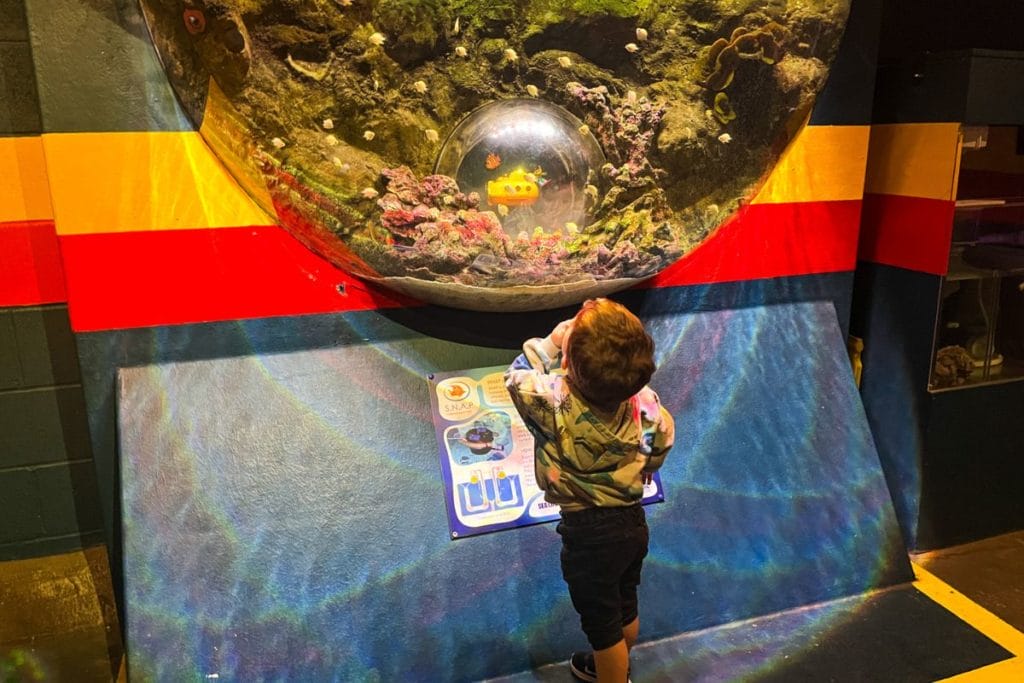 A young child marvels at a unique aquarium display shaped like a dome at a Merlin Pass aquarium, featuring colorful coral and a yellow submarine model.