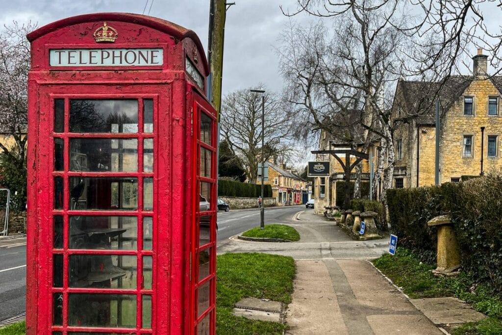 A classic red British telephone box stands on a quiet village street lined with stone buildings and leafless trees. The overcast sky adds a subdued tone to the quaint, picturesque setting.