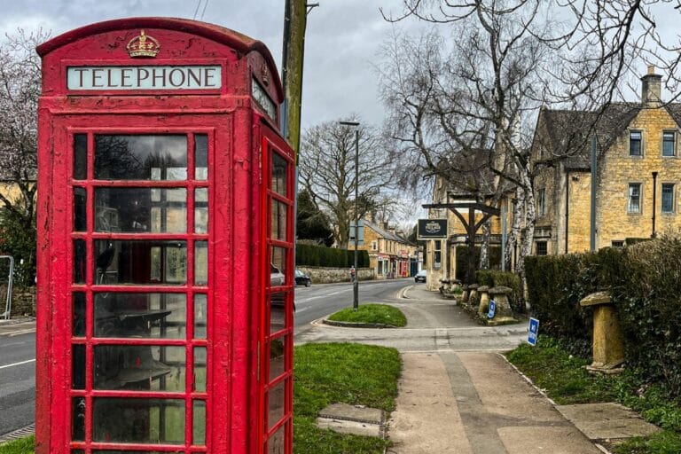 A classic red British telephone box stands on a quiet village street lined with stone buildings and leafless trees. The overcast sky adds a subdued tone to the quaint, picturesque setting.