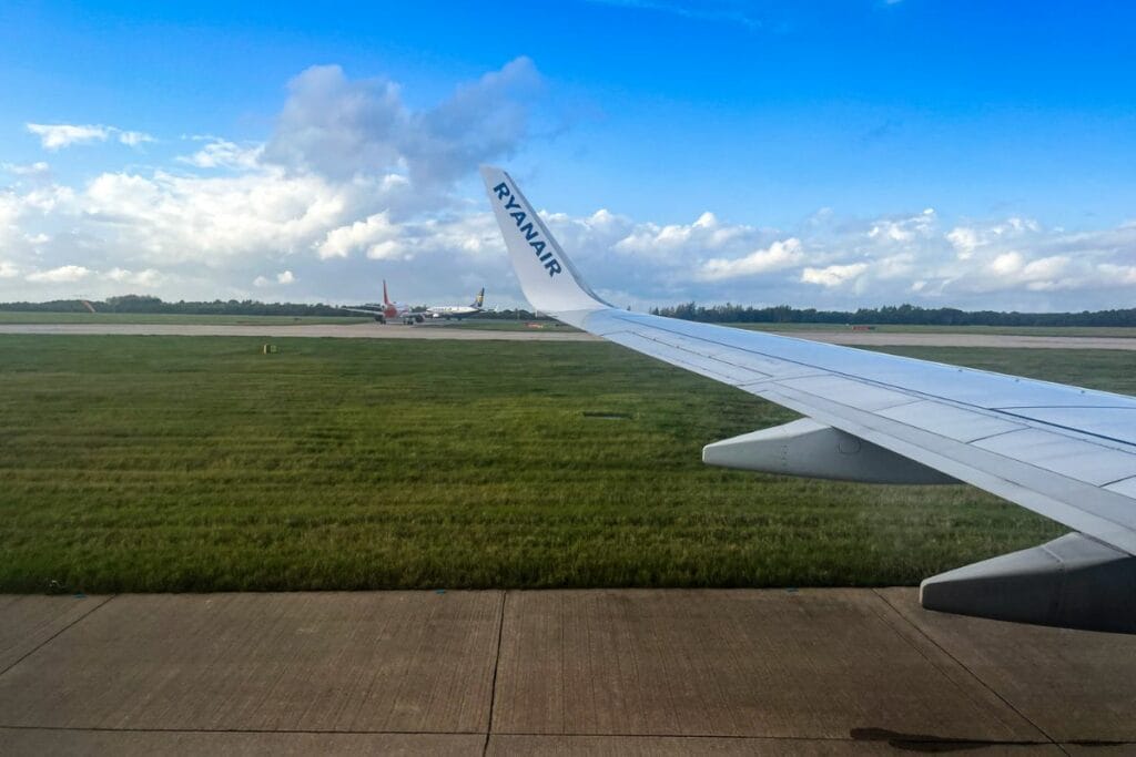 1 Airport in the UK View from a Ryanair airplane window of the airport runway. Several planes are visible in the background, and the sky is a mix of blue and clouds, creating a calm scene before takeoff.