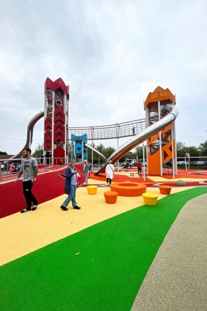 1 Butlins Sky Park Colourful playground at the Sky Park at Butlins. There are tall red and orange towers connected by a bridge and large slides. People walk and play on the vibrant ground, featuring various shapes and play structures under a cloudy sky.