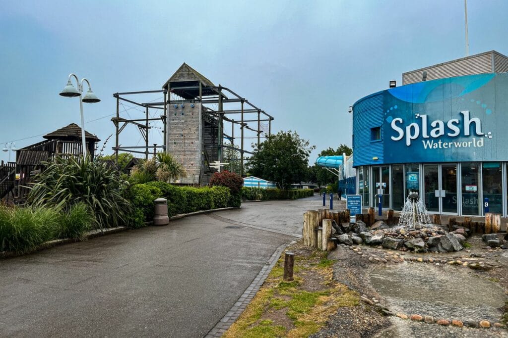 Acitvities at Butlins Minehead A wide-angle view of the entrance to Splash Waterworld at Butlins Minehead. A building with a blue façade and the Splash Waterworld sign stands prominently on the right, while an outdoor climbing structure and lush greenery line the path on the left.