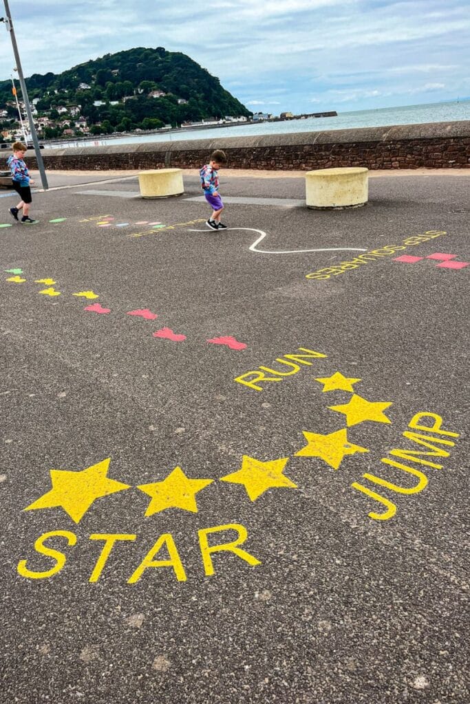Minehead Beach Walk A scenic pathway along Minehead Beach, featuring colorful markings for a children's play area labeled "Star Jump." Two children wearing matching tops play near the seawall, with a hilly landscape and ocean in the background.