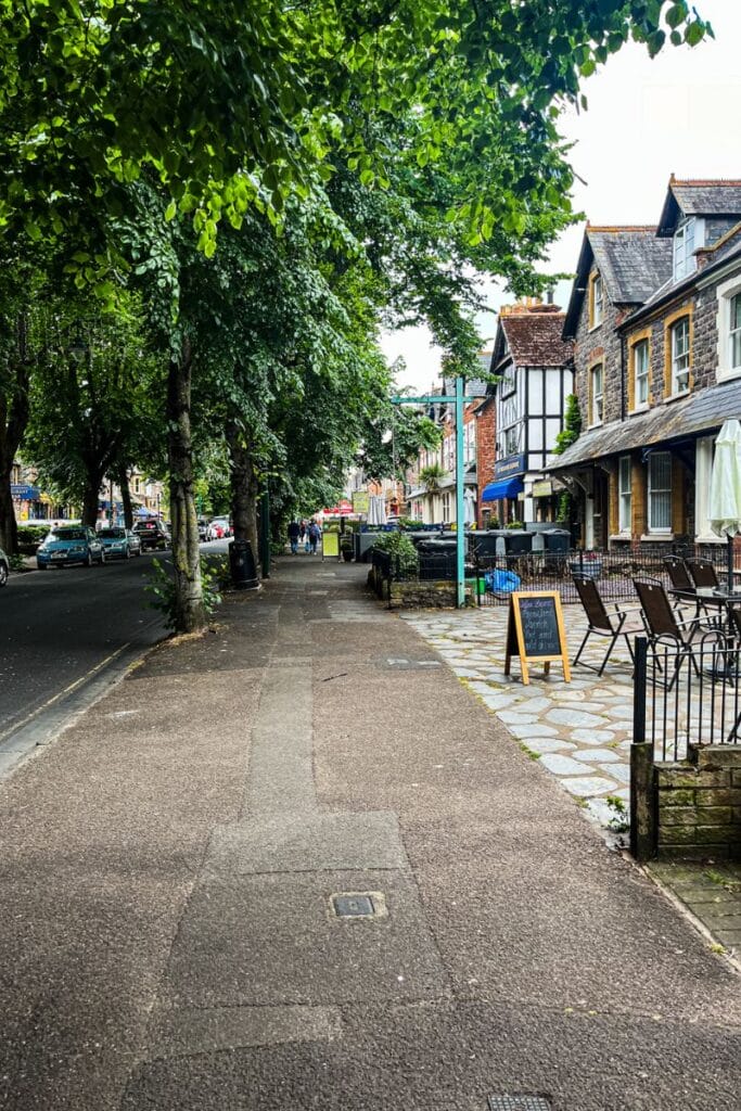 Minehead Town Centre A quaint street scene in a town center features a wide sidewalk lined with trees on the left and a row of charming old buildings on the right. Outdoor seating with chairs and tables is visible, along with a chalkboard sign advertising a menu. The street has light traffic and a few pedestrians in the distance, with a canopy of green leaves overhead providing shade.
