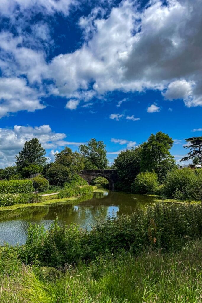 UK Countryside The image shows a tranquil countryside scene with a stone bridge arching over a calm river. The river is surrounded by lush green vegetation, and the sky above is a vibrant blue with fluffy white clouds scattered across. Tall trees line the riverbank, adding to the serene, natural setting.
