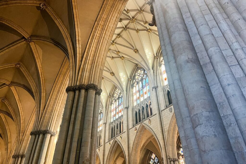 A close-up view of soaring Gothic arches and ribbed vaults of the York Minster, with sunlight streaming in through ornate stained-glass windows, emphasizing the architecture’s grandeur.