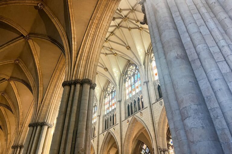 A close-up view of soaring Gothic arches and ribbed vaults of the York Minster, with sunlight streaming in through ornate stained-glass windows, emphasizing the architecture’s grandeur.