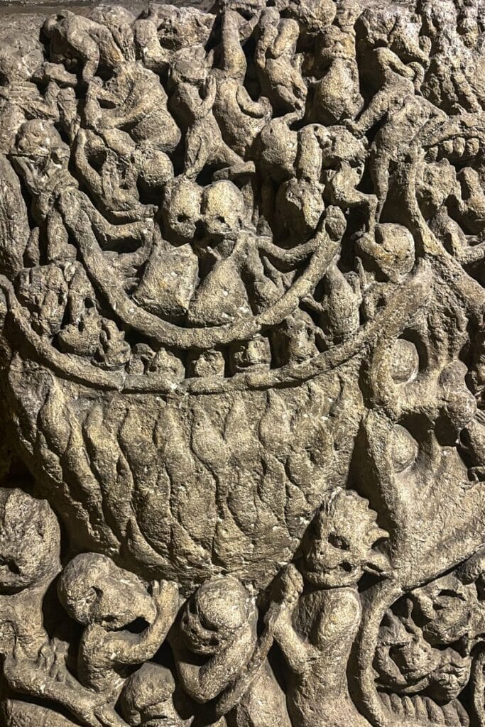 A detailed stone carving depicting souls in torment, with figures inside a cauldron, demonic creatures, and distressed human forms, possibly from a medieval church or cathedral.
