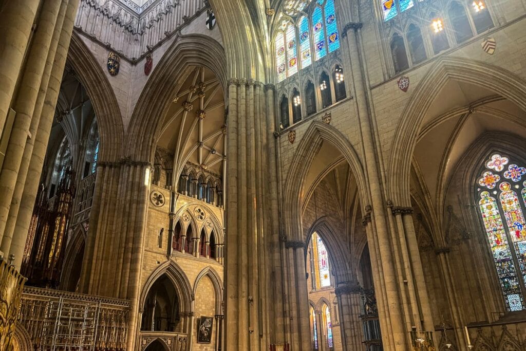 The grand interior of the Gothic cathedral, the York Minster with towering columns, pointed arches, intricate stonework, and vibrant stained-glass windows filtering light into the sacred space.